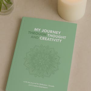 Könnte beinhalten: Ein mintgrünes Journal mit dem Text "MY JOURNEY THROUGH THOUGHT AND CREATIVITY" und einem floralen Design. Das Journal dient der Reflexion, dem Wachstum und dem kreativen Ausdruck. Eine brennende weiße Kerze und eine Vase mit weißen Blumen sind im Hintergrund.