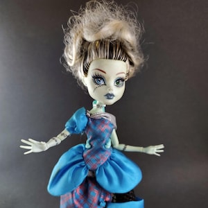 May include: A doll with pale green skin, styled blonde hair, and mismatched eyes. The doll wears a blue dress with a plaid bodice and a ruffled skirt. The doll's arms are outstretched, and it has stitched details on its arms.