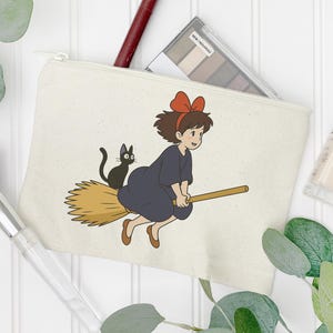 May include: A cream-colored canvas cosmetic bag featuring a cartoon illustration of a young witch riding a broom with a black cat. The witch wears a dark blue dress and a red bow.