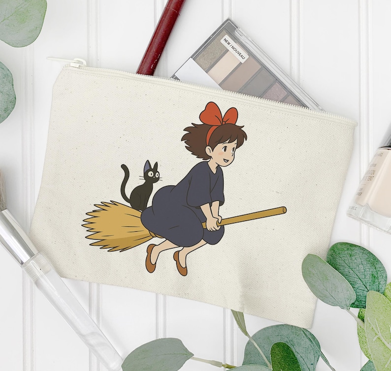 May include: A cream-colored canvas cosmetic bag featuring a cartoon illustration of a young witch riding a broom with a black cat. The witch wears a dark blue dress and a red bow.