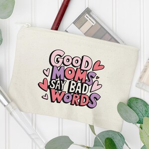 Puede incluir: Bolsa de maquillaje de lona color crema con cierre de cremallera. La bolsa presenta el texto "Good Moms Say Bad Words" en rosa, morado y negro, con detalles de corazones. Se ve una paleta de sombras de ojos y un pincel de maquillaje.