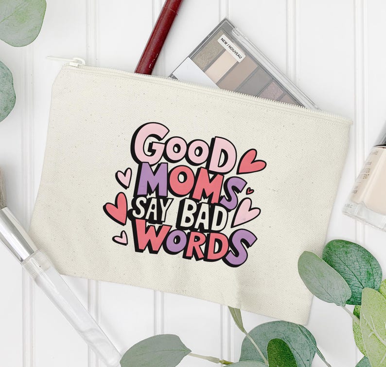 Puede incluir: Bolsa de maquillaje de lona color crema con cierre de cremallera. La bolsa presenta el texto "Good Moms Say Bad Words" en rosa, morado y negro, con detalles de corazones. Se ve una paleta de sombras de ojos y un pincel de maquillaje.