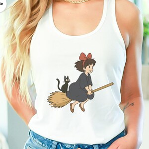 May include: White tank top featuring a cartoon illustration of a young witch riding a broom with a black cat. The witch wears a dark gray dress and a red bow. The broom is light brown.