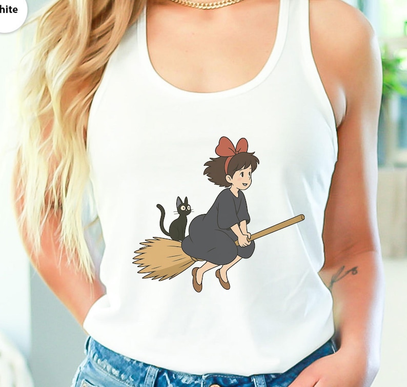 May include: White tank top featuring a cartoon illustration of a young witch riding a broom with a black cat. The witch wears a dark gray dress and a red bow. The broom is light brown.