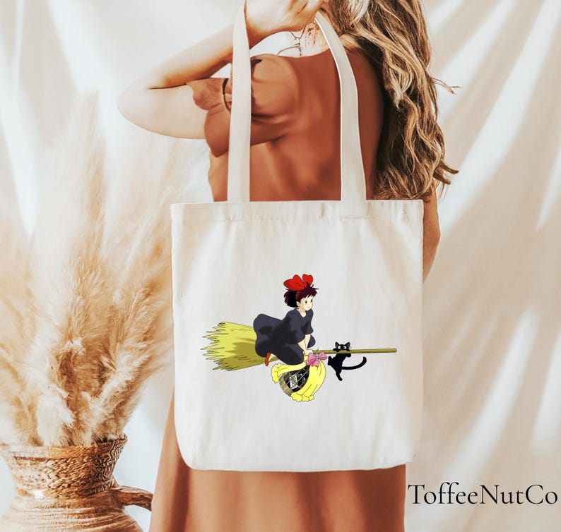 May include: A white tote bag featuring a cartoon illustration of a young witch riding a broom with a black cat. The witch is wearing a black dress and a red bow. The broom has a yellow straw. The bag has cream-colored handles.