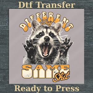 Peut inclure: Un transfert DTF avec un dessin de raton laveur, la bouche ouverte et les pattes levées. Le texte "DIFFERENT Day SAME Shit" est en orange et blanc, avec "Ready to Press" en dessous. Le raton laveur est en noir et blanc sur fond gris.