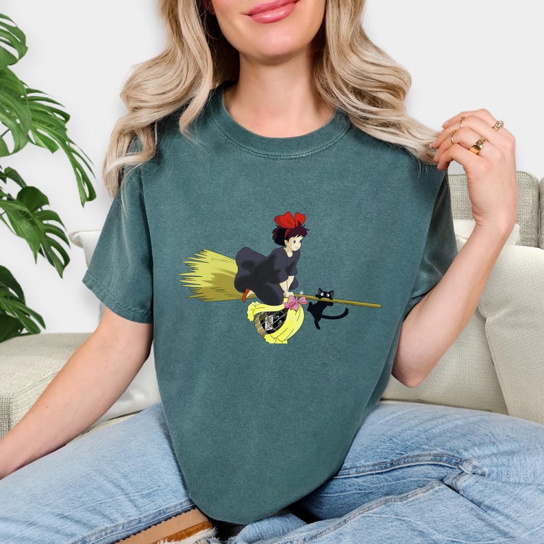 May include: Teal-colored t-shirt featuring a cartoon illustration of a witch riding a broom with a black cat. The witch wears a black dress and a red bow. The broom is yellow and the cat is black.