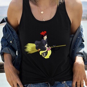 May include: Black tank top featuring a cartoon illustration of a young witch riding a broom. The witch wears a black dress and a red bow. A black cat sits on the broom. The broom is yellow and the witch is flying over a yellow bag.