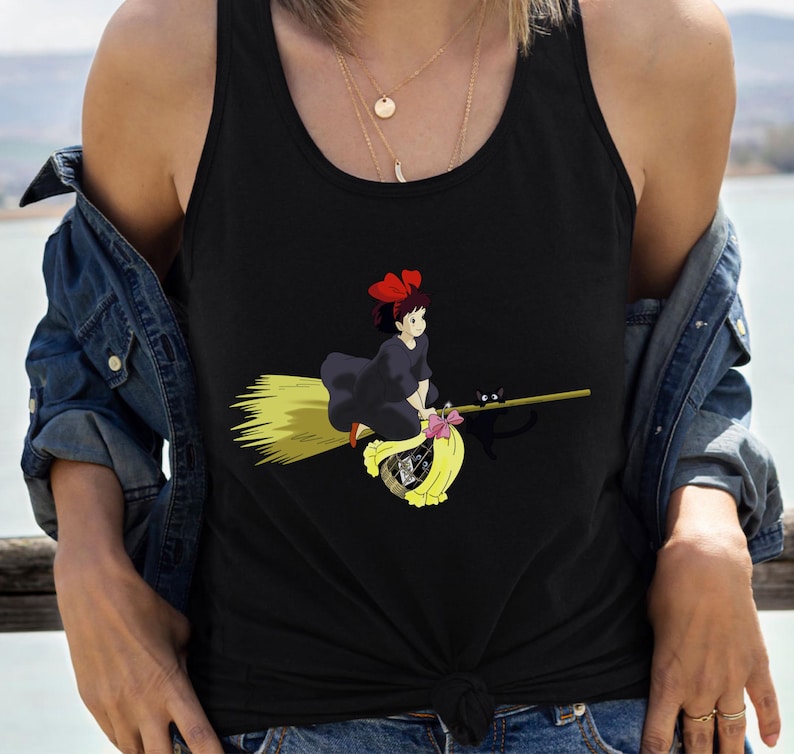 May include: Black tank top featuring a cartoon illustration of a young witch riding a broom. The witch wears a black dress and a red bow. A black cat sits on the broom. The broom is yellow and the witch is flying over a yellow bag.