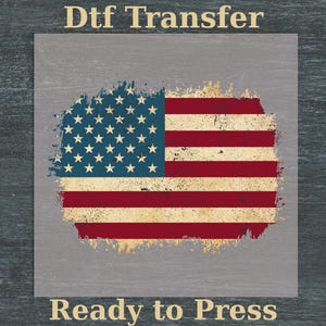 May include: A distressed American flag design with red and white stripes and blue stars. The words "DTF Transfer" and "Ready to Press" are printed in a gold color above and below the flag image. The background is a gray square.