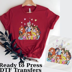 以下が含まれることがあります： 様々なプリンセスが描かれたカラフルなグラフィックが特徴の赤いTシャツ。デザインにはお城、ミッキーマウスの形をした風船、そして下部には「Ready to Press DTF Transfers」の文字が含まれています。木製のハンガーにかけられています。