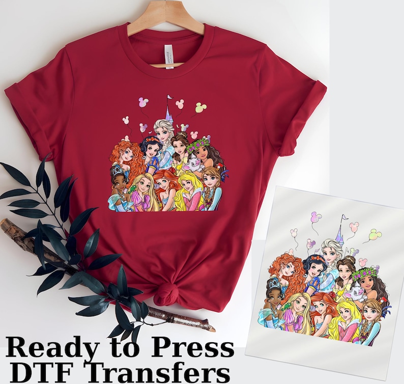 以下が含まれることがあります： 様々なプリンセスが描かれたカラフルなグラフィックが特徴の赤いTシャツ。デザインにはお城、ミッキーマウスの形をした風船、そして下部には「Ready to Press DTF Transfers」の文字が含まれています。木製のハンガーにかけられています。