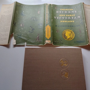 May include: An open book titled "Charles Dickens and Early Victorian England" by R.J. Cruikshank. The cover is green with gold lettering and illustrations. The book is displayed with its pages open, revealing text and illustrations.