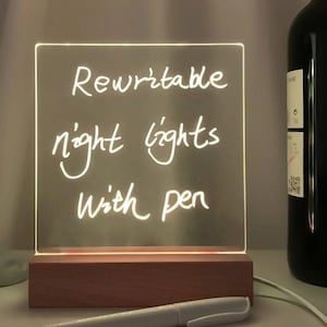 May include: A clear acrylic square sign with a wooden base, lit from behind, with the words "Rewritable night lights with pen" written on it in white marker. A white marker pen is lying on a table in front of the sign.