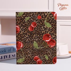 May include: A notebook with a leopard print background and red cherry illustrations. The notebook has a white border and the text "Piyana Gifts" in the upper right corner. The cherries have green leaves.
