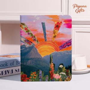 Puede incluir: Un diario con un diseño de puesta de sol colorido. La portada presenta una vibrante puesta de sol con rayos naranjas, amarillos y morados. El fondo incluye montañas, flores y el texto "Piyana Gifts".