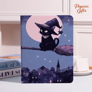 May include: A notebook featuring a black cat wearing a witch hat, riding a broomstick against a night sky backdrop. The design includes a full moon, stars, and a cityscape. The text "Piyana Gifts" is in the upper right corner.