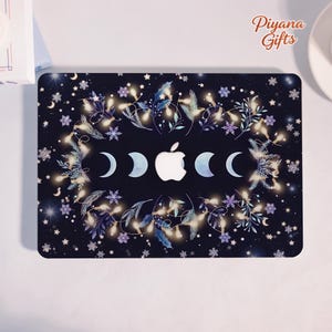 May include: A black laptop skin featuring a central white Apple logo surrounded by a horizontal sequence of moon phases. The design includes glowing string lights, blue foliage, and white snowflakes on a dark background with the text Piyana Gifts.