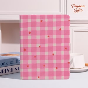 May include: A pink and white checkered notebook with red star accents. The notebook is standing upright, with the text "Piyana Gifts" in the upper right corner. A book titled "Book of Br" is visible in the background.