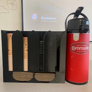 May include: Black coffee cup holder with three compartments for cups and two compartments for lids. The holder is next to a red thermos with the words "Family Owned Community Coffee" printed on it.