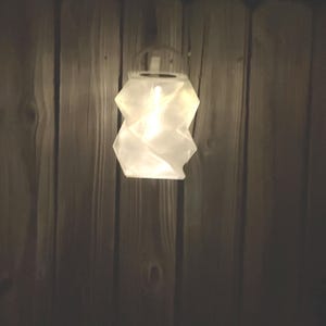 Könnte beinhalten: Eine leuchtende, weiße Laterne mit geometrischem Design hängt vor einem Holzzaun. Das Licht der Laterne beleuchtet ihre gefaltete, papierartige Struktur und erzeugt eine warme Atmosphäre. Der Hintergrund ist ein dunkler, vertikaler Holzzaun.