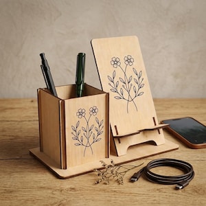 Puede incluir: Un organizador de escritorio de madera clara que cuenta con un portalápices cuadrado y un soporte para teléfono integrado. Ambos componentes están decorados con una ilustración de líneas negras de dos flores con hojas. Un bolígrafo negro y uno verde están dentro.