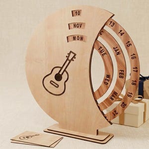 Puede incluir: Un calendario perpetuo de madera clara con un gráfico de guitarra, que cuenta con anillos circulares giratorios que muestran meses, días de la semana y fechas. El texto incluye 10, NOV, MON, TUE, WED, JAN, FEB, MAR y los números del 13 al 21.