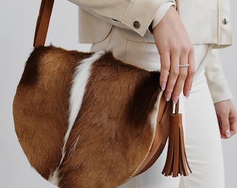 May include: A brown and white cowhide handbag with a long brown strap and a brown tassel. The bag is worn over the shoulder of a person wearing a white jacket and white pants.