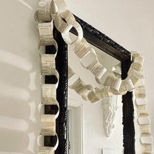 May include: A decorative paper chain garland crafted from vintage book pages with visible printed text, draped over a rectangular mirror featuring a dark, ornate, carved frame. A metallic star ornament is mounted on the wall to the right side.