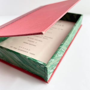 Könnte beinhalten: Geöffnete rechteckige Box mit rotem Deckel und grün marmorierten Seiten. Im Inneren befindet sich gedruckter Text auf cremefarbenem Papier. Die Box dient wahrscheinlich zur Aufbewahrung von Büchern oder anderen kleinen Gegenständen. Die Box ist aus Pappe.