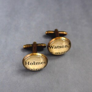 May include: A pair of vintage-style cufflinks with a bronze finish. Each cufflink has a clear, domed glass over a printed background with the names "Holmes" and "Watson" in black text. The cufflinks are set against a muted blue background.