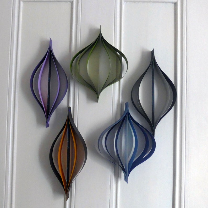 May include: Five paper ornaments in various colors, including purple, green, blue, and orange, are hanging on a white wall. The ornaments are made of paper strips that are folded and layered to create a 3D effect.