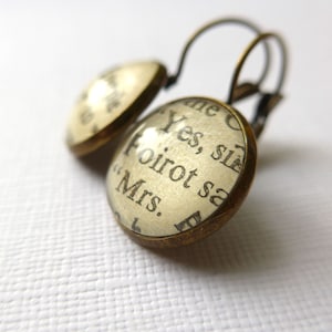 May include: A pair of gold-toned earrings with a round glass charm featuring a vintage book page with the text "Yes, sir, Poirot said "Mrs."