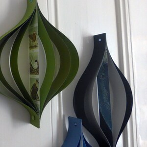 May include: Three paper ornaments in the shape of a teardrop. The ornament on the left is green and white with a bird illustration. The ornament in the center is black and white. The ornament on the right is blue and white.