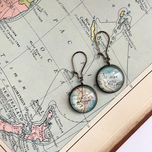 May include: A pair of antique brass-toned earrings. Each earring features a round, glass-covered map design with the words "St Ives" and "Lerwick" visible. The earrings are set against a vintage map background.