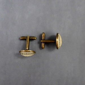 May include: A pair of gold-toned cufflinks with a white background. The cufflinks feature the text "Holmes" and "Watson" in black.