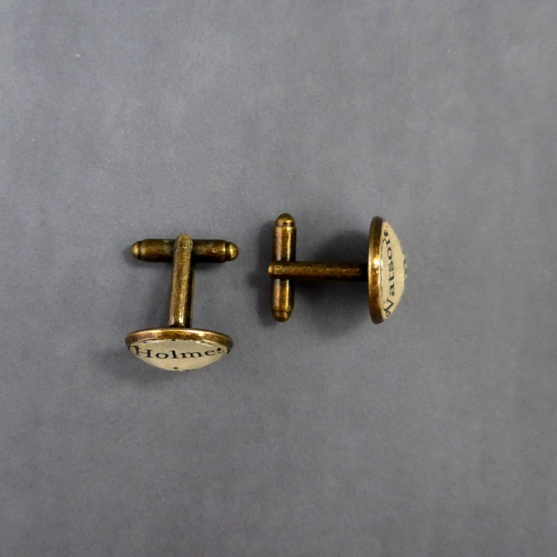 May include: A pair of gold-toned cufflinks with a white background. The cufflinks feature the text "Holmes" and "Watson" in black.