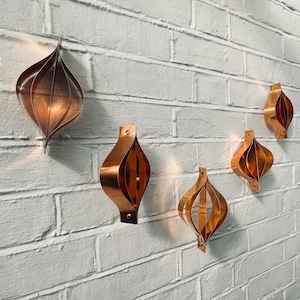 May include: Copper-colored, geometric ornaments strung on a wire, hanging against a white brick wall. The ornaments have a teardrop shape with curved, layered metal. The warm tones of the copper contrast with the cool white of the wall.