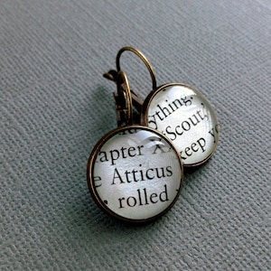 May include: A pair of silver hoop earrings with glass charms featuring text from the book "To Kill a Mockingbird". The charms read "Chapter X" and "Atticus rolled" on one earring and "...ything Scout, keep yo" on the other.