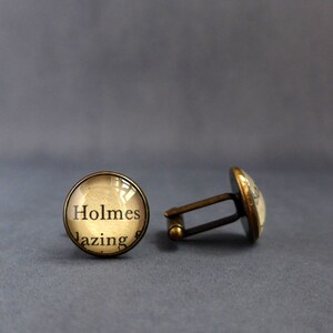 May include: A pair of gold-toned cufflinks with a round glass design. The glass features a vintage book page with the text "Holmes lazing".