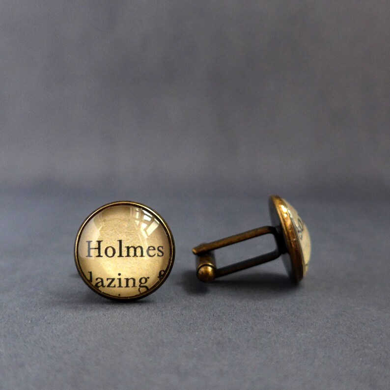May include: A pair of gold-toned cufflinks with a round glass design. The glass features a vintage book page with the text "Holmes lazing".