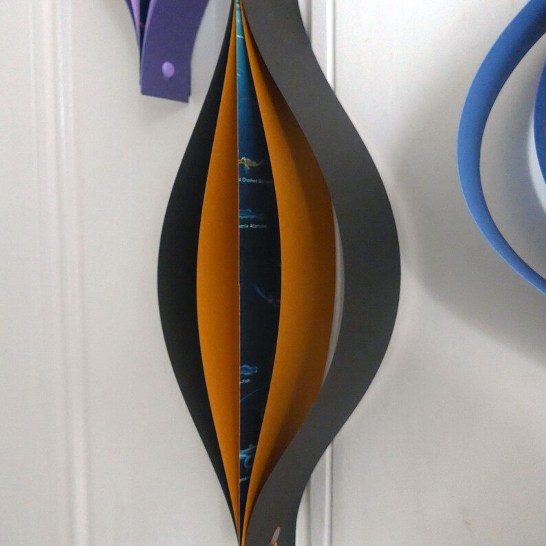 May include: A black and orange paper ornament with a folded design. The ornament is hanging from a white wall.