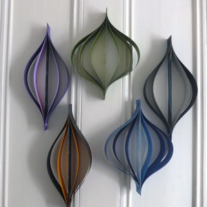 May include: Five paper ornaments in different colors, including purple, green, gray, blue, and orange. The ornaments are hanging on a white wall.