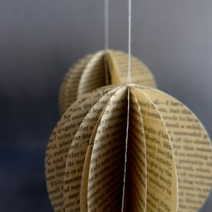 May include: A paper ornament made from pages of a book. The ornament is white and beige and has a starburst shape. It is hanging from a white string.