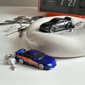 May include: Two miniature car keyrings. One is a sleek black sports car, and the other is a blue car with orange accents. The blue car keyring has a white dice and a silver charm attached. Both keyrings are on a white surface.