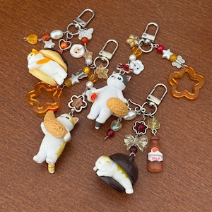 May include: Four keychains featuring white cat figurines paired with various charms. The cats are depicted with food items like bread and pastries. The keychains include silver-toned lobster clasps, colorful beads, star-shaped charms, and a small brown bottle labeled Strawberry Milk.