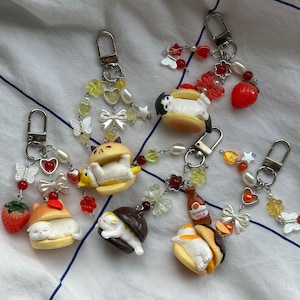 May include: A selection of keyrings with small cat figures inside food items. Each keyring has a silver clasp and is decorated with beads, bows, and charms, including strawberries and hearts. The cats are white and cream-coloured.