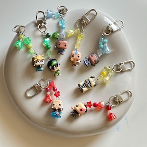 Puede incluir: Una colección de ocho llaveros con figuras en miniatura unidas a mosquetones plateados. Cada llavero está decorado con cuentas de plástico de colores en formas de flores, estrellas y corazones, mostrando diversos diseños de personajes.