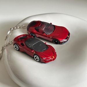 Op de afbeelding: Twee metallic rode Ferrari speelgoedauto sleutelhangers met gedetailleerde zwarte interieurs, getinte ramen en zilverkleurige metalen kettingen. Elk miniatuurvoertuig toont het iconische gele Ferrari-embleem op de motorkap.