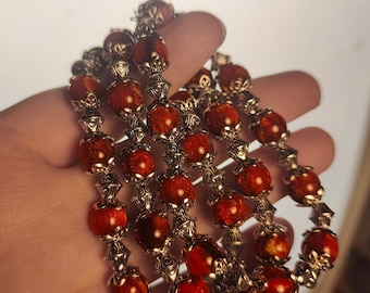 May include: A necklace featuring round, translucent orange-red beads with a swirling, marbled texture. Each bead is separated by intricate, silver-toned metal filigree caps that add a decorative, textured element to the strand of beads.